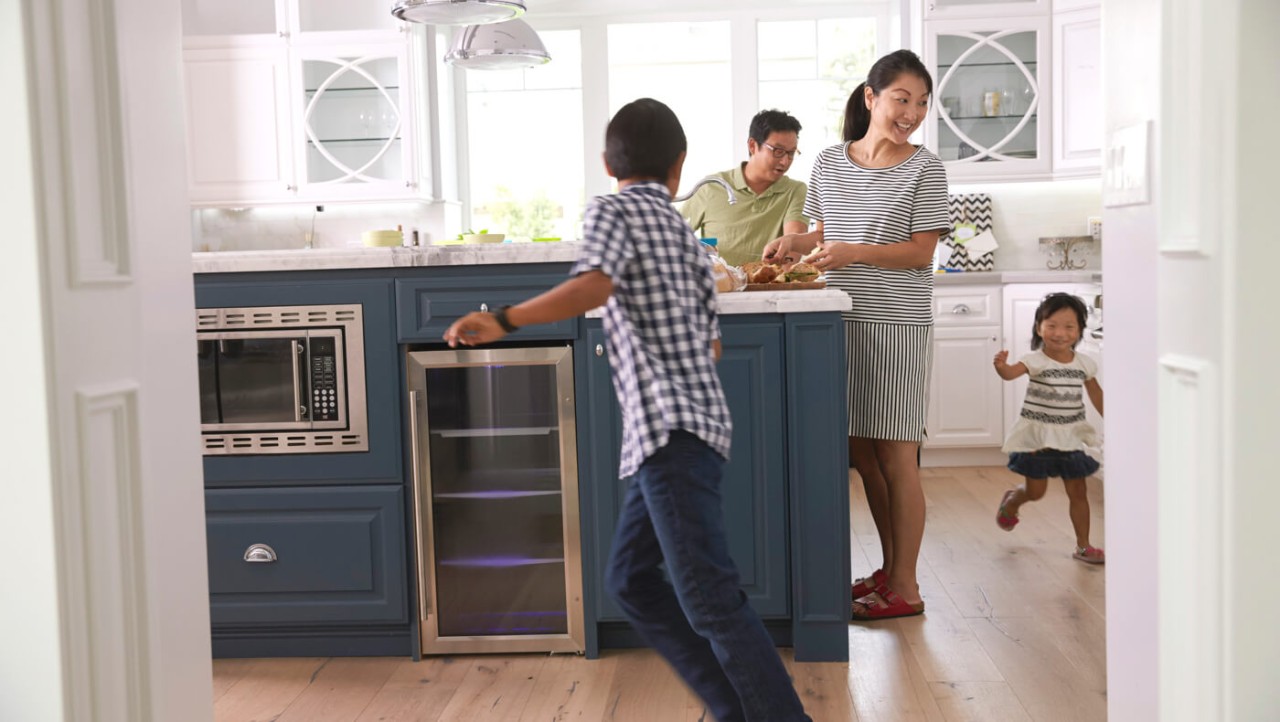 30-something parents watch two young children chase each other in their kitchen.