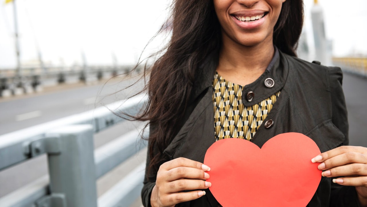 A young woman smiles and holds a red paper heart.