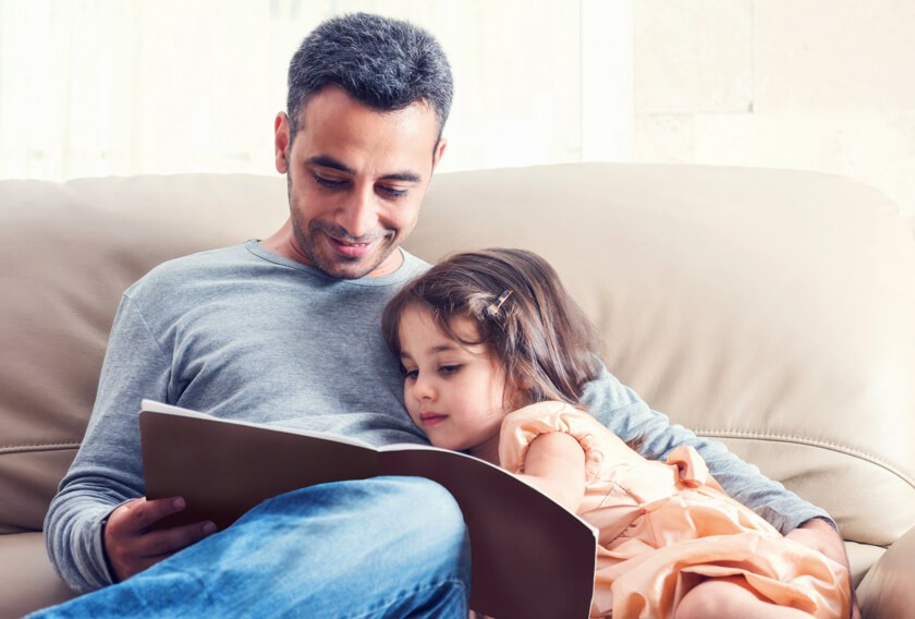 A smiling 30-something dad reads to his young daughter.