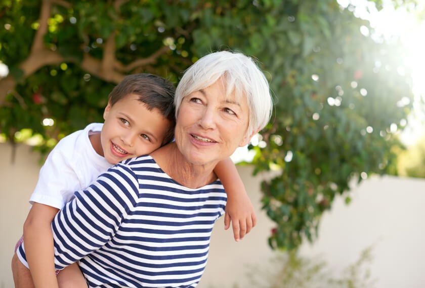 An older smiling woman carries her grandson on her back.